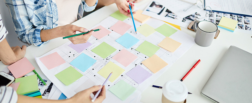 Mapeamento do processo de vendas: colaboradores apontam canetas para diferentes post its coloridos na mesa