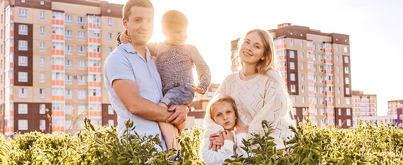 Marketing de Experiência: Família composta por homem, mulher e duas crianças sorriem ao ar livre para a câmera com um conjunto de imóveis ao fundo