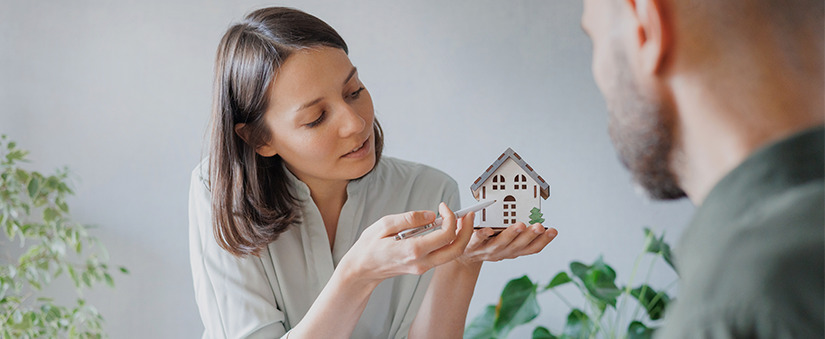 Especulação imobiliária: mulher segura maquete de casa em 3D. Homem olha a peça.