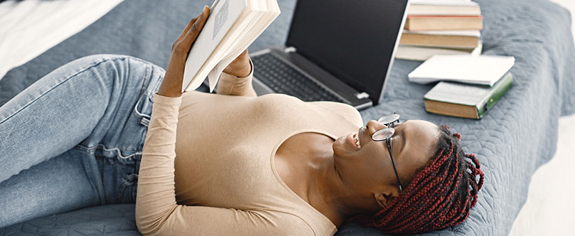 gestão de tempo e produtividade: mulher lendo deitada ao lado de um notebook e alguns livros.
