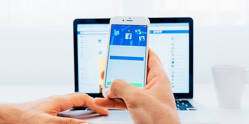 gerenciador de anúncio do facebook: imagem de uma mão segurando o celular na frente de um notebook com a página de login do facebook aberta.