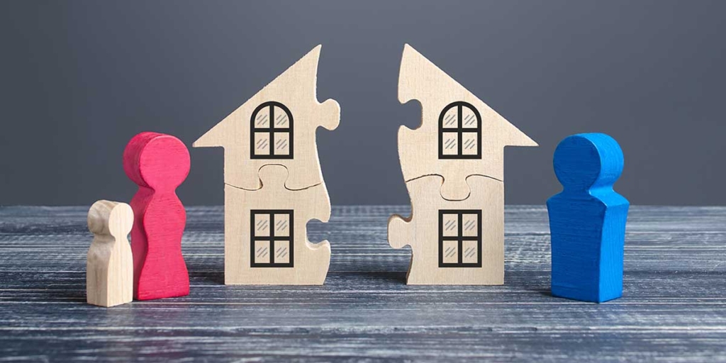 O que é permuta de imóveis: imagem de uma casa de madeira dividida ao meio e, ao lado de cada parte, bonecos de madeira nas cores rosa e azul.