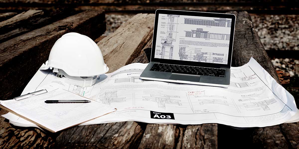 Notícias da Construção Civil: mesa com um notebook, um capacete de proteção e uma planta de edifício.