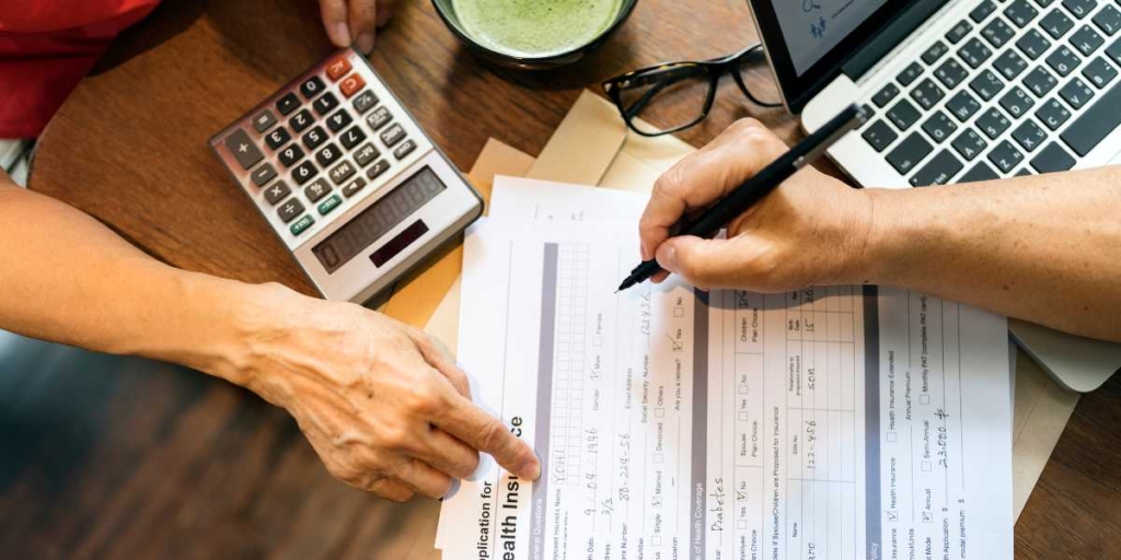 Repasse de financiamento imobiliário: imagem de uma mesa vista de cima com alguns papeis, uma calculadora, um notebook e mãos de duas pessoas em cima de um papel.
