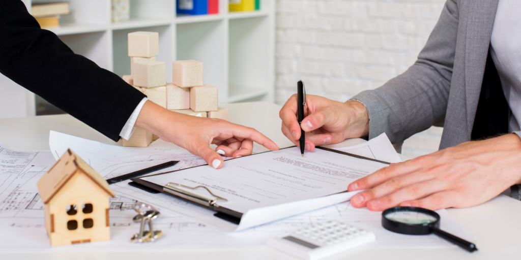 Agente imobiliário ou Corretor de imóveis: close em mãos de pessoas de lados opostos de uma mesa. Uma delas assina um papel e a outra aponta para o documento.