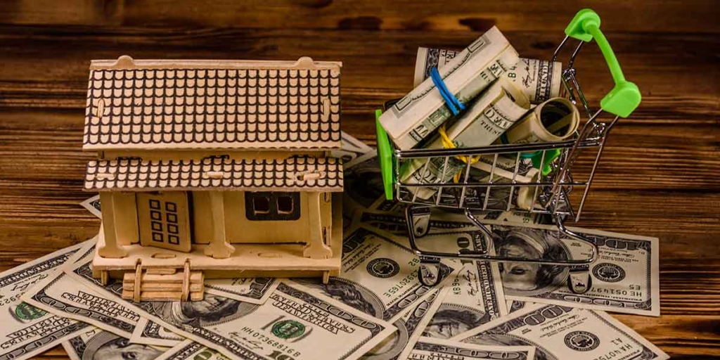 Área de Corretagem: imagem de uma miniatura de carrinho de compras carregando várias cédulas de dinheiro. Ao lado uma casa de madeira. 