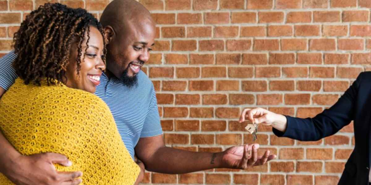 Cliente 4.0: homem e mulher negros se abraçam e sorriem enquanto o homem estende a mão para pegar um molho de chaves de uma corretora de imóveis branca.
