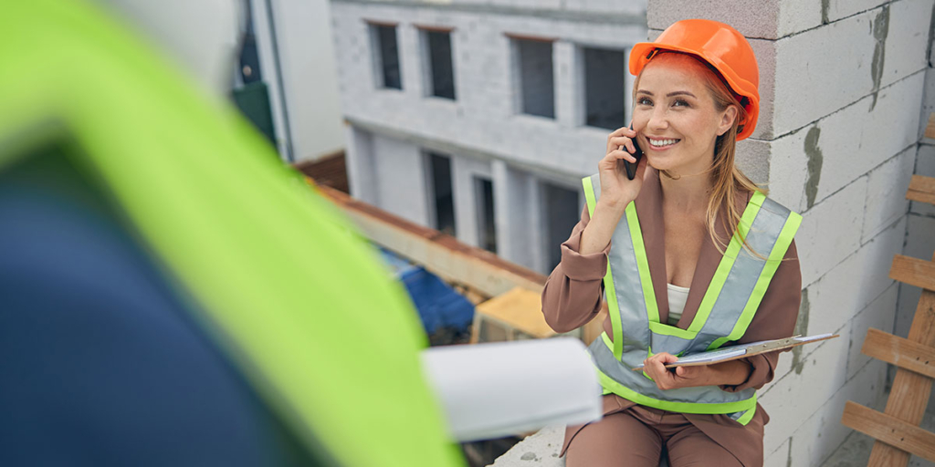 Atendimento ao cliente na construção civil: imagem de uma mulher usando EPIs num canteiro de obras, sorrindo, segurando um celular próximo ao ouvido.