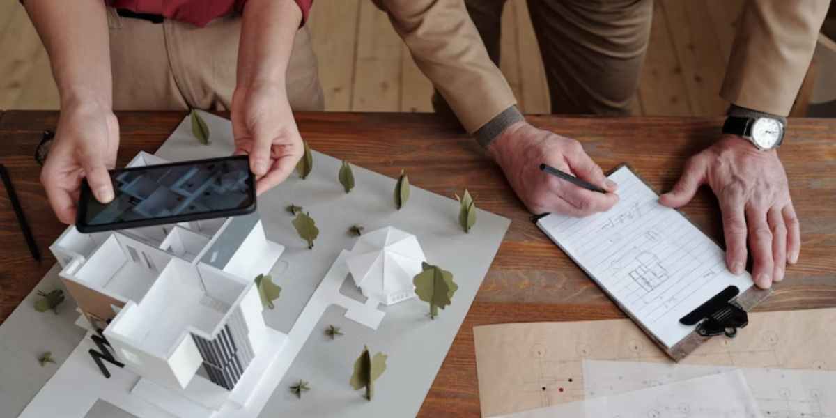 Avaliação de imóveis: imagem de uma mesa vista de cima, com uma maquete de edifício e as mãos de uma pessoa segurando um celular e tirando uma foto. Do lado esquerdo, mãos de outra pessoa escrevendo num papel.