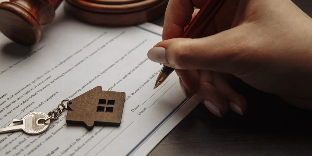 Documento de venda de imóvel: imagem de uma mão assinando um papel. Ao lado, uma chave com chaveiro em formato de casa.