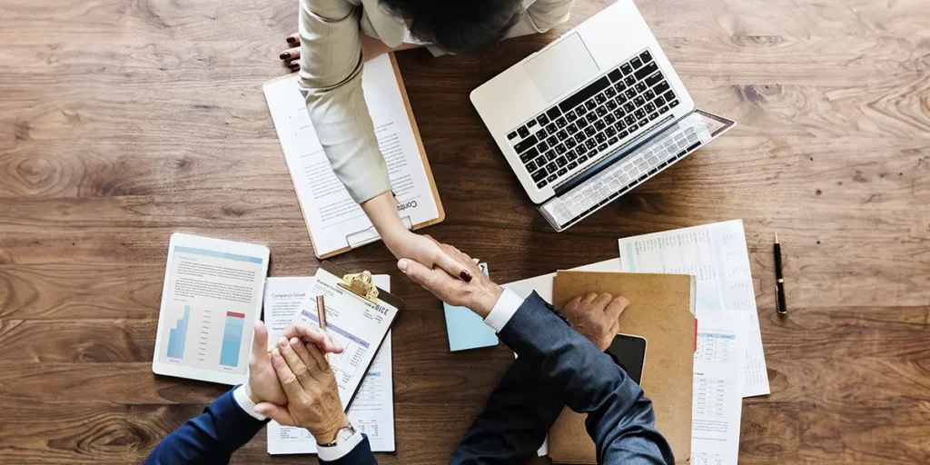 Mediação Imobiliária: imagem de uma mesa vista de cima com alguns papeis, notebook e mãos de três pessoas. Duas delas apertam as mãos.