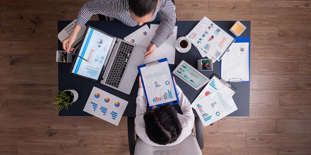 Quais são os tipos de CRM: mesa vista de cima com duas mulheres sentadas à frente. Na mesa vários papeis com gráficos coloridos.