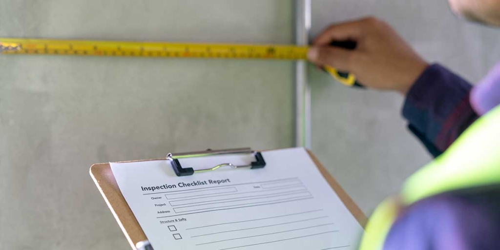 Relatório de visita técnica: close numa pessoas segurando um papel com o título "Inspection checklist report". Com a outra mão, ela mede o comprimento de uma parede.