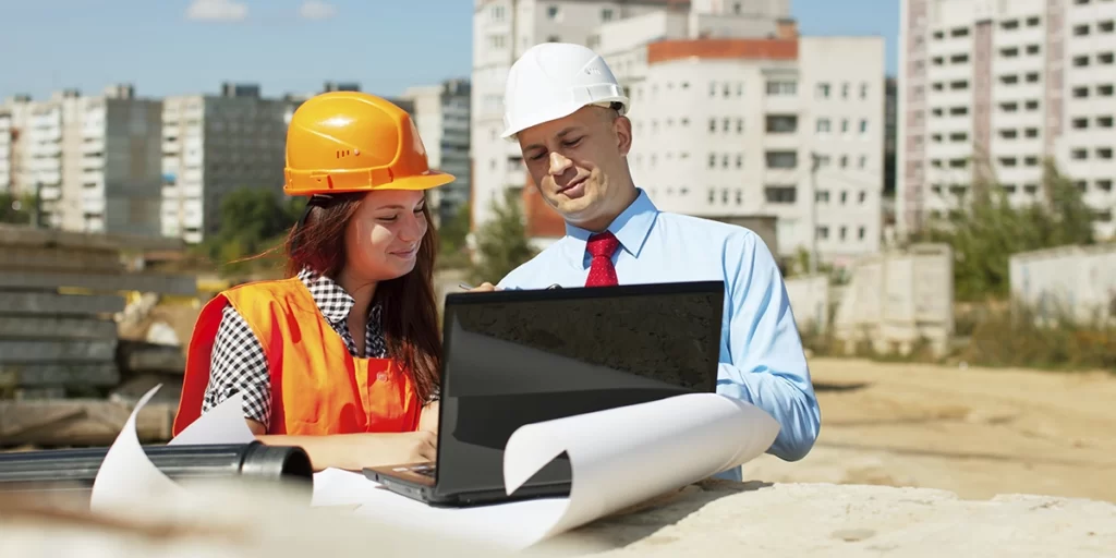 ABNT da Construção Civil: imagem de um homem e uma mulher conversando num canteiro de obras. Os dois usam capacete e estão olhando para a tela de um notebook.