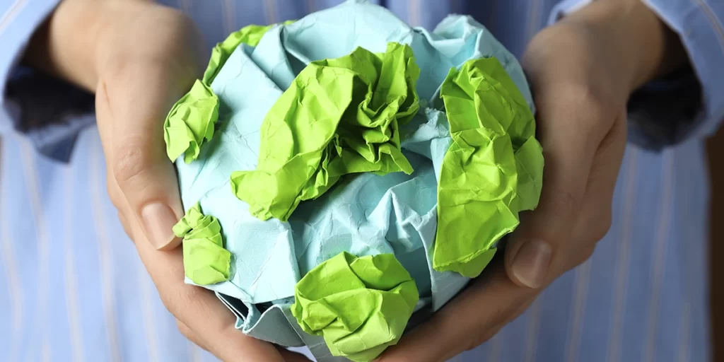 Materiais Ecológicos: close nas mãos de uma pessoa segurando um papel azul amassado com alguns detalhes verdes em alusão ao planeta Terra.