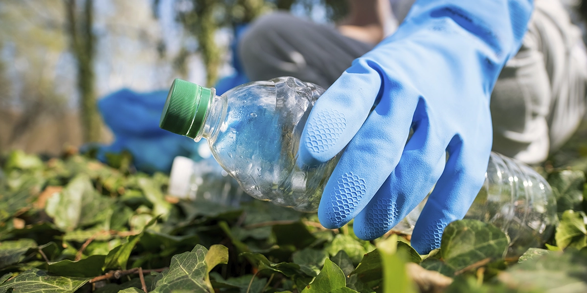 Materiais Ecológicos: close nas mãos de pessoas que usam luvas, seguram garrafas de plástico e jogam o líquido da garrafa em plantas.