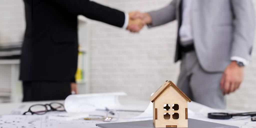 Negócios imobiliários: imagem de uma mesa cheia de papeis. Em destaque, uma casa de madeira em miniatura. Ao fundo dois homens apertando as mãos.