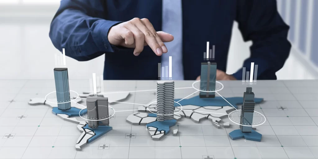 PropCo: close na mão de uma pessoa atrás de uma mesa apontando para a ilustração em 3D de um edifício na mesa. Em cima da mesa vemos ilustrações de um mapa e alguns prédios. 