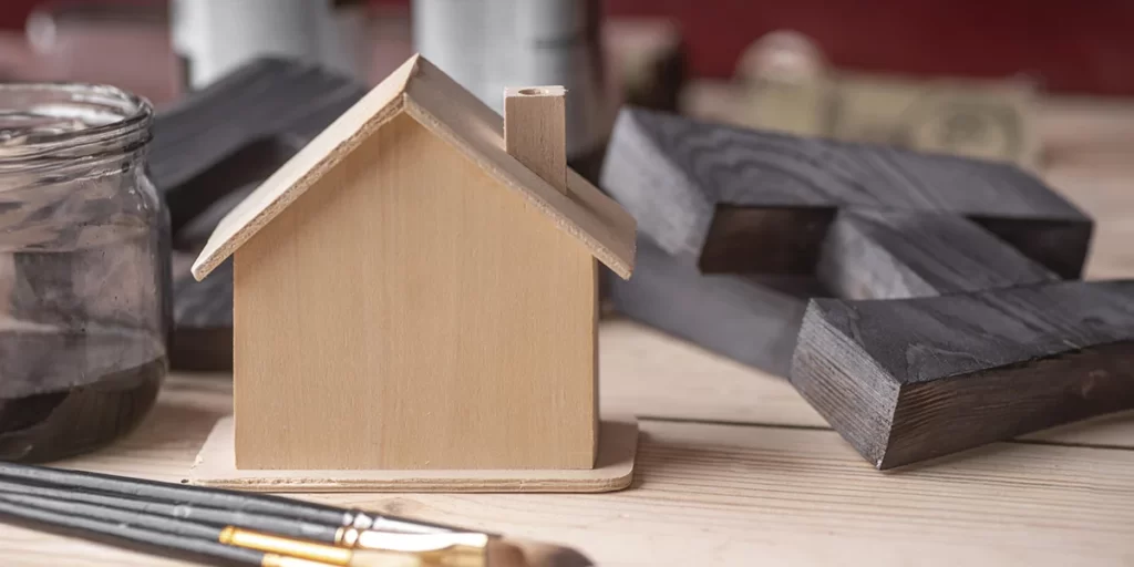 Casas Pré-fabricadas: imagem de uma mesa com algumas peças de madeira e pinceis de tinta. Em destaque, a miniatura de uma casa de madeira.