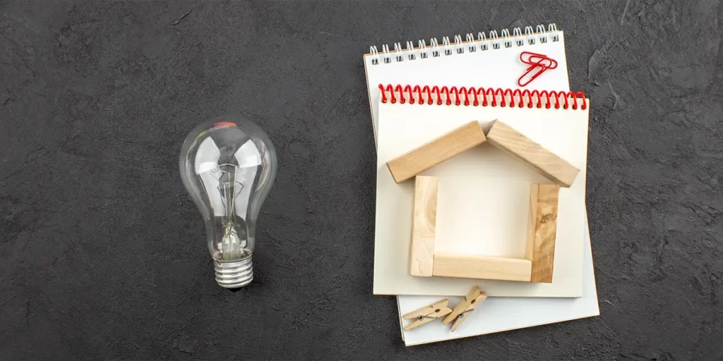 Casas Pré-fabricadas: imagem de uma lâmpada numa superfície plana. Ao lado da lâmpada, o contorno de uma casa de madeira em cima de alguns cadernos em branco.