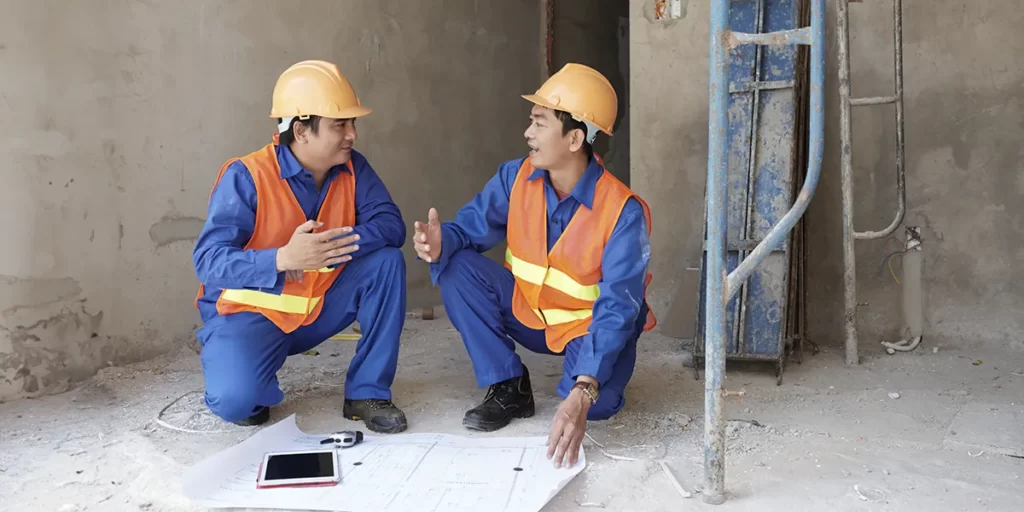 Como calcular custo de obra: dois homens fardados, usando capacetes estão num canteiro de obras. Os dois estão agachados e conversam.