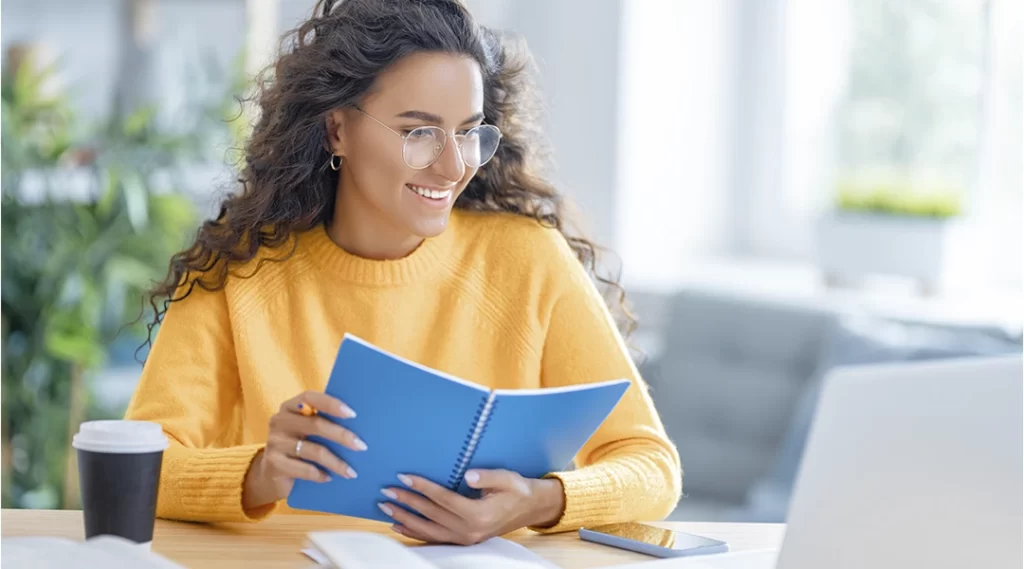 Cursos para corretor de imóveis: imagem de uma mulher sorridente, sentada à frente de uma mesa. Ela segura um caderno azul e olha para um notebook.