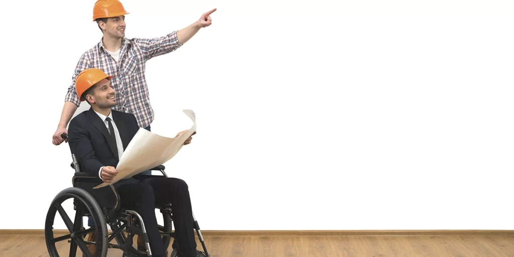Norma de Acessibilidade: imagem de dois homens usando capacete de proteção. Um deles está num cadeira de rodas, segurando uma planta de imóvel. Os dois sorriem e olham para o mesmo lugar.