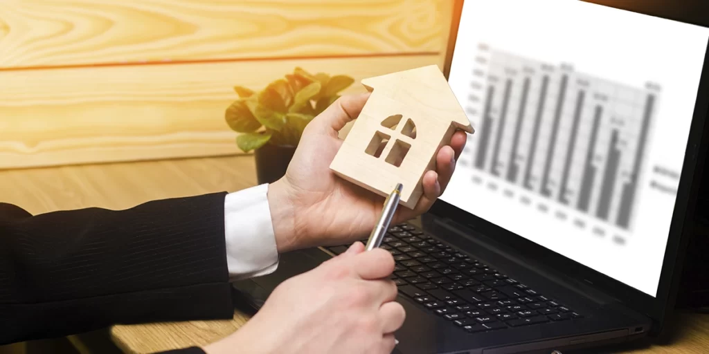 Portifólios imobiliários: close nas mãos de uma pessoa segurando uma miniatura de casa de madeira. Ao fundo um notebook com um gráfico na tela.