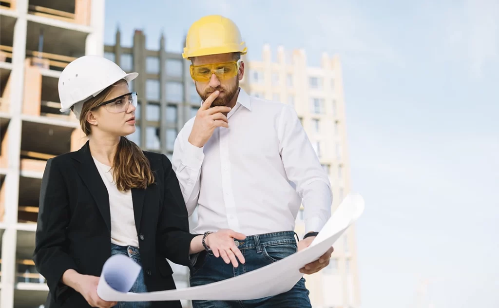 Administração de Construtora: imagem de um homem e uma mulher num canteiro de obras, usando EPI. Eles seguram um papel grande e conversam apontando para ele.  