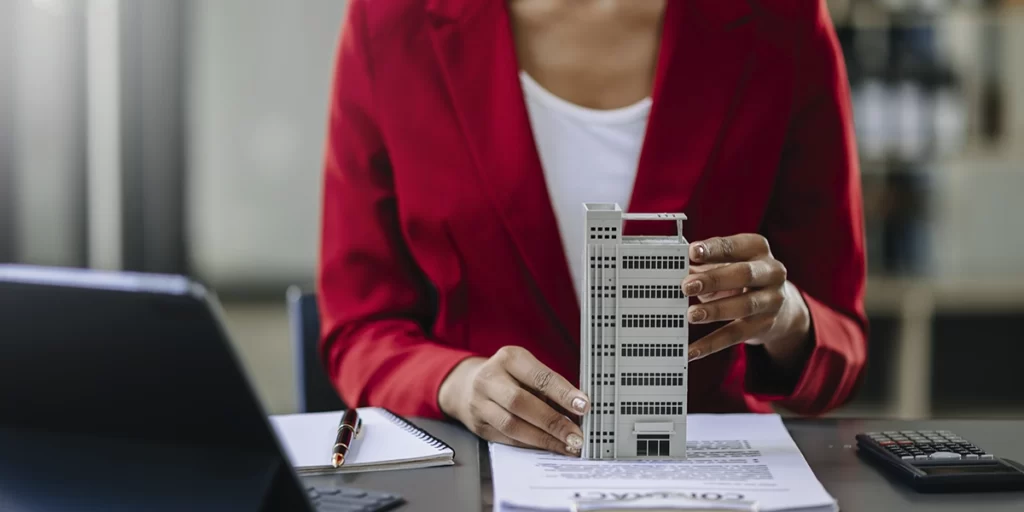 Administração de Imóveis: imagem das mãos de uma mulher segurando uma miniatura de edifício em cima de uma mesa de escritório.