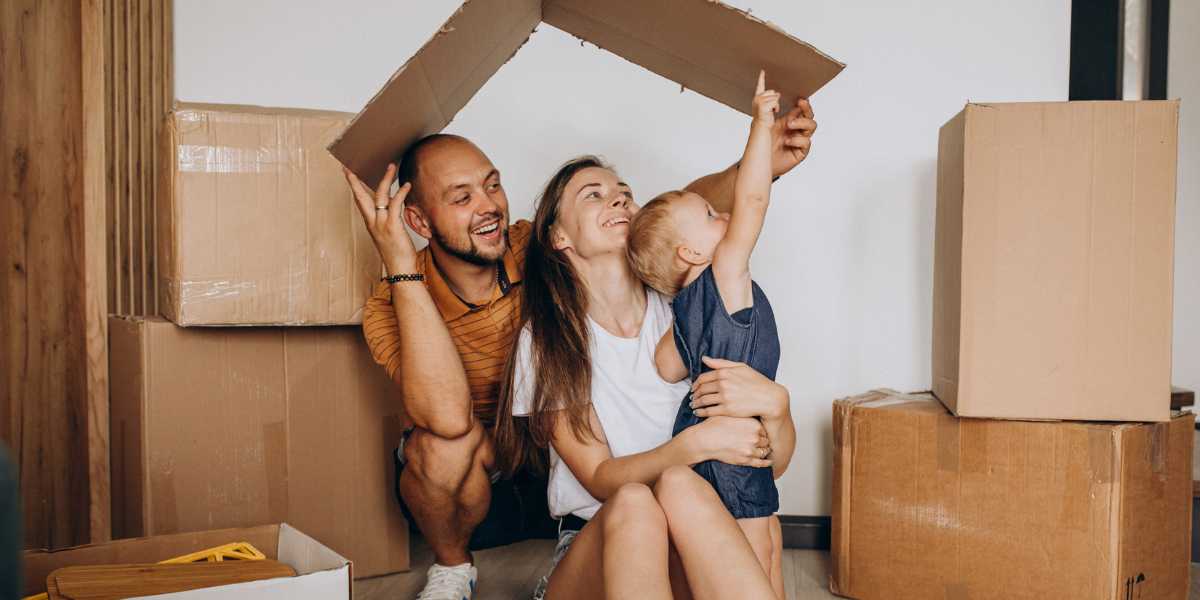 Novas regras do minha casa minha vida: imagem de uma família (um homem, uma mulher e uma criança) brincando com caixas de papelão, formando um telhado.