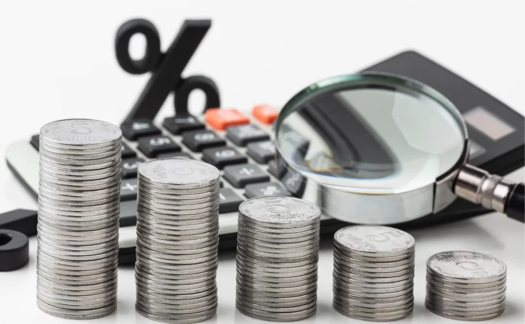 Taxa SELIC: imagem de pilhas de moeda de tamanhos diferentes, da maior pilha para a menor (esquerda para direita). Ao fundo uma calculadora e uma lupa.