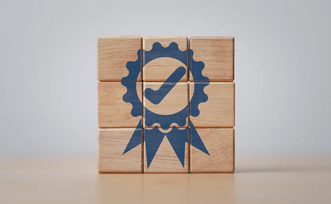 gestão da qualidade na construção civil: A imagem mostra um cubo de madeira com um logotipo em azul e branco. O logotipo parece ser um símbolo de qualidade ou certificação, com um círculo e um símbolo de verificação. O cubo está sobre um fundo neutro, possivelmente um ambiente de escritório ou uma superfície de trabalho.