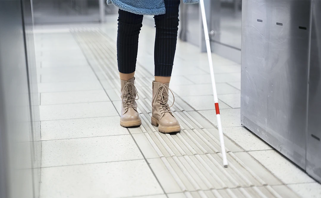 nbr 9050 atualizada: A imagem mostra uma pessoa usando botas e caminhando com o auxílio de um guia para deficientes visuais. A imagem é tirada de uma angulação baixa e a pessoa está se aproximando da câmera.