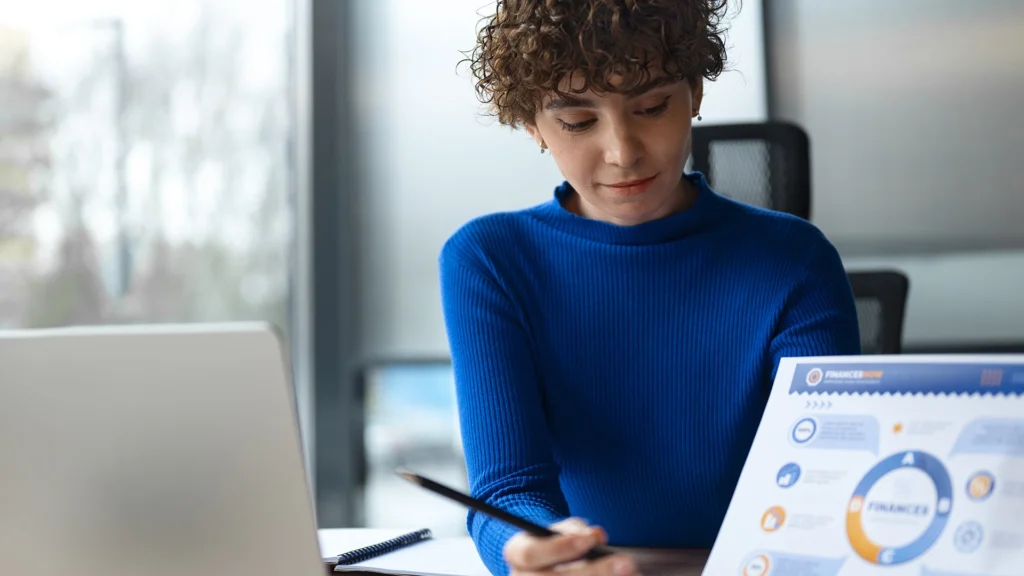 CRM reduz custos: A imagem mostra uma pessoa sentada em uma mesa, focada em um documento ou plano de negócios que está em frente a ela. A pessoa está vestindo uma blusa azul e parece estar pensativa ou concentrada em algo. Ela tem cabelos encaracolados e está usando um colar. Ao lado, há um laptop aberto, sugerindo que ela pode estar trabalhando ou estudando. Ao fundo, há uma janela com uma vista de edifícios, indicando que a cena pode estar acontecendo em um ambiente de escritório ou de trabalho.