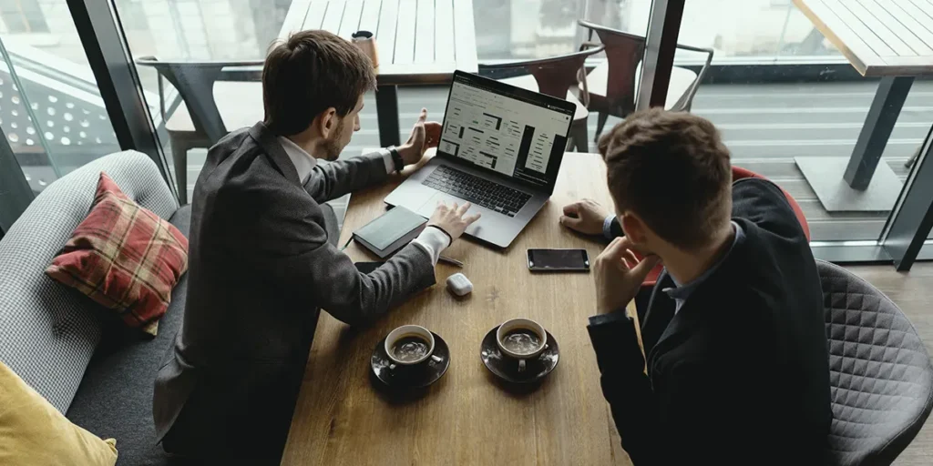 Como vender Imóveis para investidores:  foto de um ângulo alto. Dois homens de terno estão sentado ao redor de uma mesa de madeira. Os dois olham para um notebook. Em cima da mesa vemos duas xícaras de café. O ambiente sugere uma cafeteria.