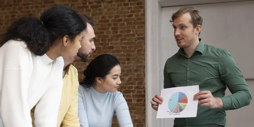 incorporadora imobiliária: A imagem mostra um grupo de cinco pessoas em um ambiente que parece ser um escritório ou sala de reunião. Um homem está de pé, segurando uma folha de papel com um gráfico circular, que parece ser um histograma ou um gráfico de barras. Ele está falando e parece estar explicando o conteúdo do gráfico para as outras pessoas. As outras quatro pessoas estão sentadas e estão olhando para ele, sugerindo que estão participando de uma discussão ou uma apresentação. Todos parecem estar vestidos casualmente e estão em um ambiente informal. A sala tem uma parede de ladrilhos e uma porta ao fundo.