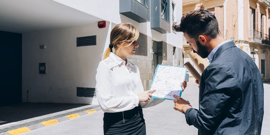 incorporadora imobiliária: A imagem mostra dois indivíduos em um ambiente urbano. A pessoa à esquerda é uma mulher com cabelo loiro, vestindo uma camisa branca e calça preta. Ela está segurando um mapa aberto e parece estar discutindo ou explicando algo ao mapa com a outra pessoa. A pessoa à direita é um homem com cabelo curto e barba, vestindo um terno preto e uma camisa azul. Ele está segurando um rolo de papel e parece estar escutando ou discutindo o conteúdo do mapa. Ambos estão de pé e parecem estar em um ambiente ao ar livre, possivelmente em frente a uma edifício, pois há uma porta aberta ao fundo. A iluminação sugere que é um dia claro.