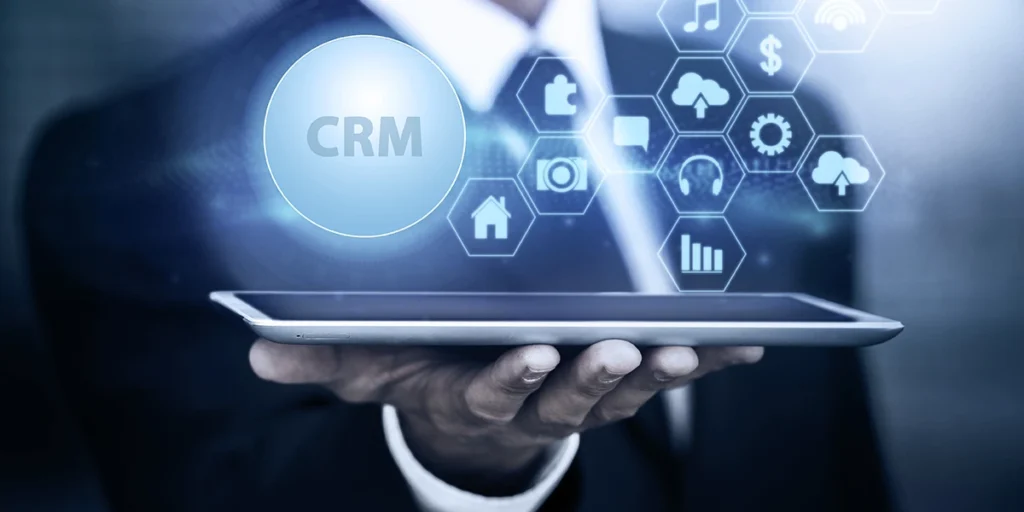 CRM para incorporadoras: close na mão de uma pessoa segurando um tablet. Da tela do tablet saem alguns ícones que representam conexão e relacionamento. Em destaque a sigla CRM.