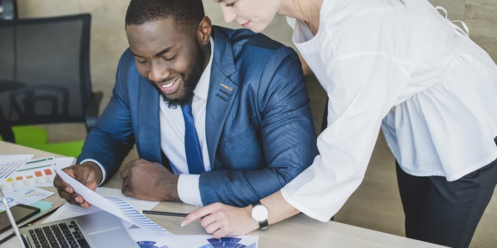 crm para gestão de incorporadora: imagem de duas pessoas num escritório. Um deles está sentado em frente a uma mesa. A outra está encostando as mãos na mesa e apontando para um papel.