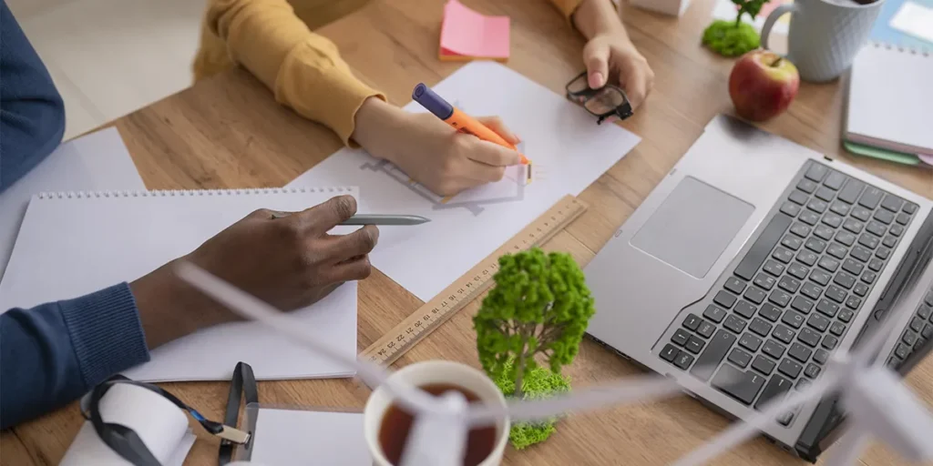 o que torna uma construção sustentável: imagem vista de cima de uma mesa de escritório com vários papeis, miniaturas de árvores e notebook. Vemos as mãos de duas pessoas que estão apoiadas na mesa com lápis e caneta na mão.