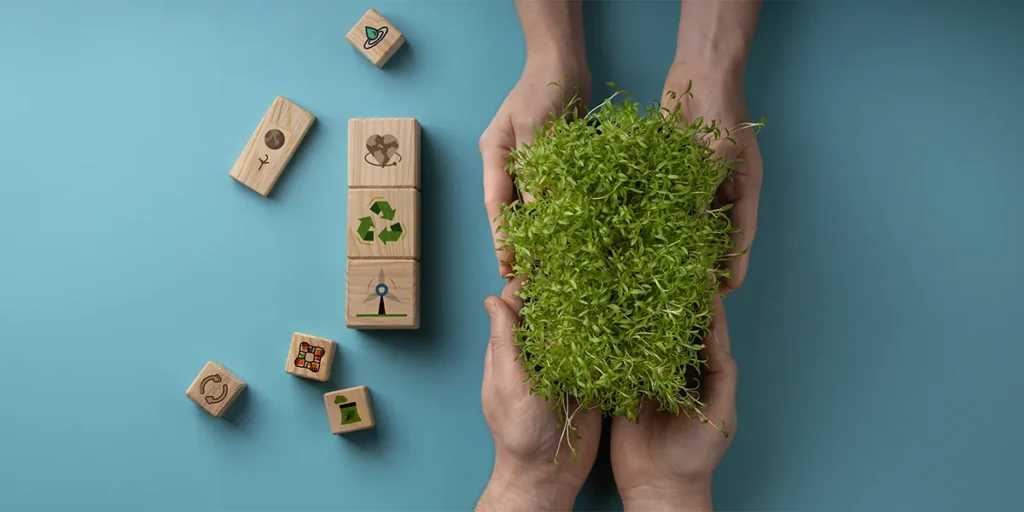 o que torna uma construção sustentável: imagem vista de cima de mãos de duas pessoas segurando algumas plantas. Ao lado, alguns blocos de madeira com ícones que representam sustentabilidade.