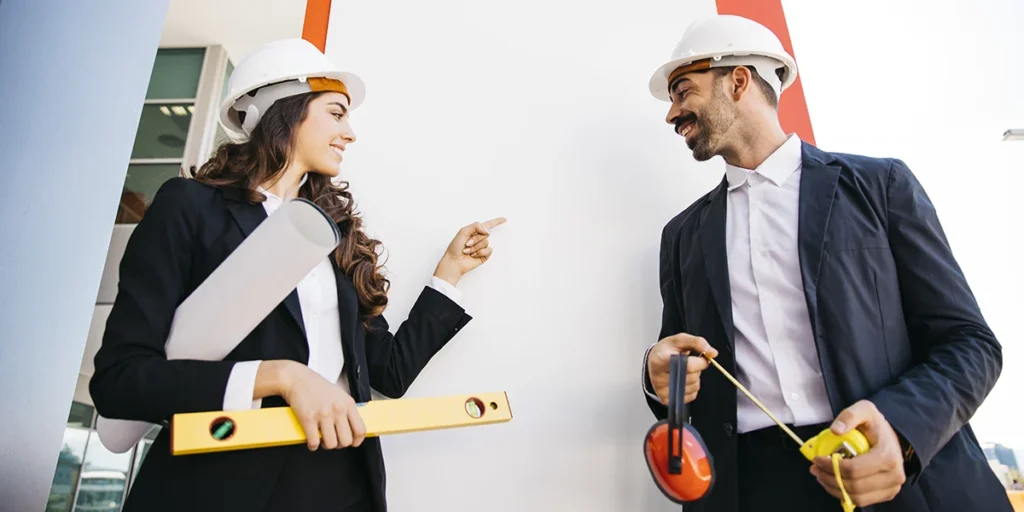  segmentação de clientes de construtoras: imagem de um homem e uma mulher conversando. Os dois usam ternos, capacete de proteção e seguram alguns instrumentos que sugere que eles estão num canteiro de obras.