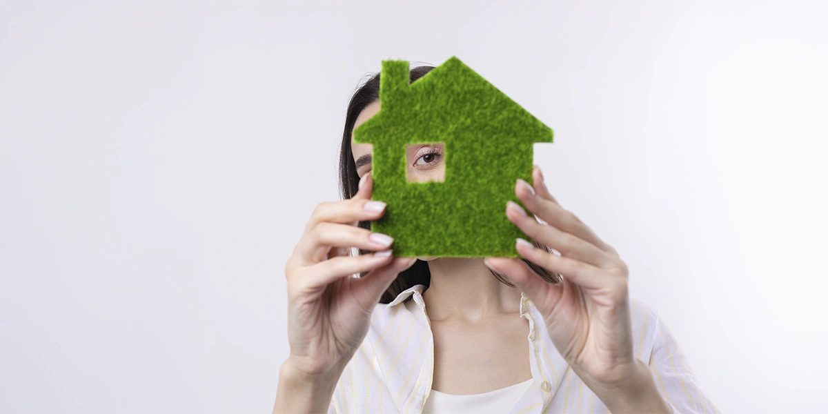 tendências do mercado imobiliário: imagem de uma mulher segurando uma miniatura de casa com um buraco para a janela. A mulher está olhando para a câmera através desse buraco.