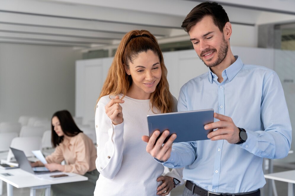 A imagem apresenta um homem e uma mulher olhando para um tablet que está nas mãos do homem. Ao fundo, uma mulher está trabalhando em seu notebook. 