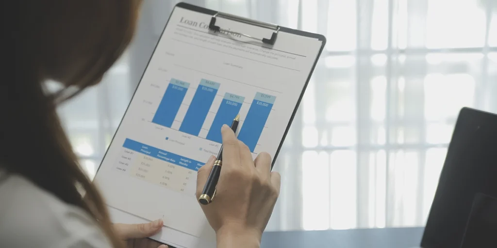 CRM para loteadoras: A imagem mostra uma mulher segurando um caderno com uma gráfica no itálico. Ela está sentada em uma mesa com um computador e um lápis na mão. O fundo é uma janela com cortinas aberto e uma vista da paisagem de uma cidade. A imagem está bem iluminada e as cores são vibrantes e vivas. A atmosfera geral da imagem é profissional e concentrada.