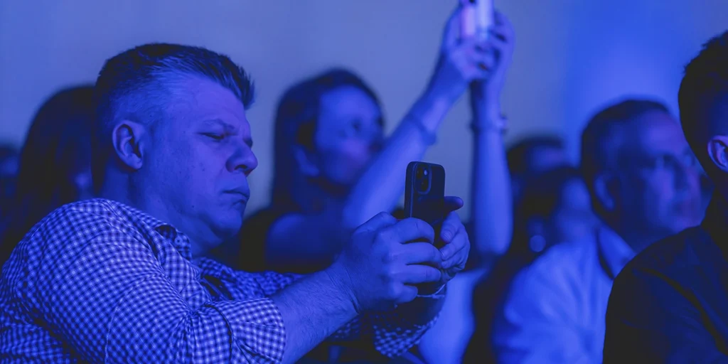 O que é o Cnstrusummit: a imagem apresenta algumas pessoas em um ambiente com luz azul. Duas delas aparecem com seus celulares nas mãos, aparentemente fazendo um vídeo ou fotografando o ambiente.