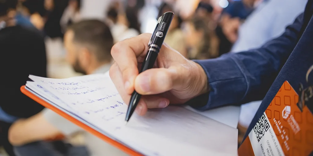 O que é o Construsummit: a imagem apresenta a mão de uma pessoa escrevendo em um caderno com uma caneta preta. Na caneta está escrito o nome do evento Construsummit, assim como no crachá na roupa da pessoa.