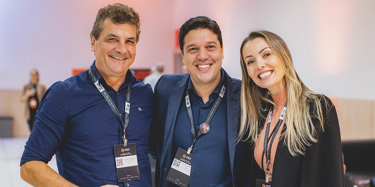 O que é o Construsummit: A imagem apresenta a foto de 3 pessoas sorrindo, sendo dois homens e uma mulher. Os três estão com crachás e aparecem em ambiente formal.