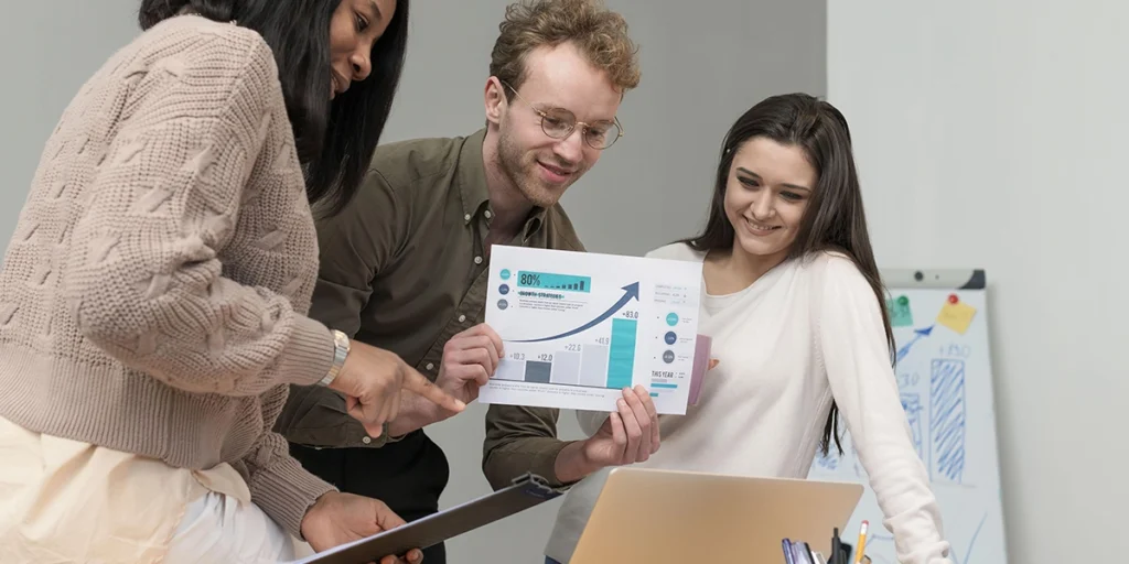 A foto possui duas mulheres e um homem durante uma reunião virtual. O homem está apresentando um gráfico em frente à tela de um notebook. 